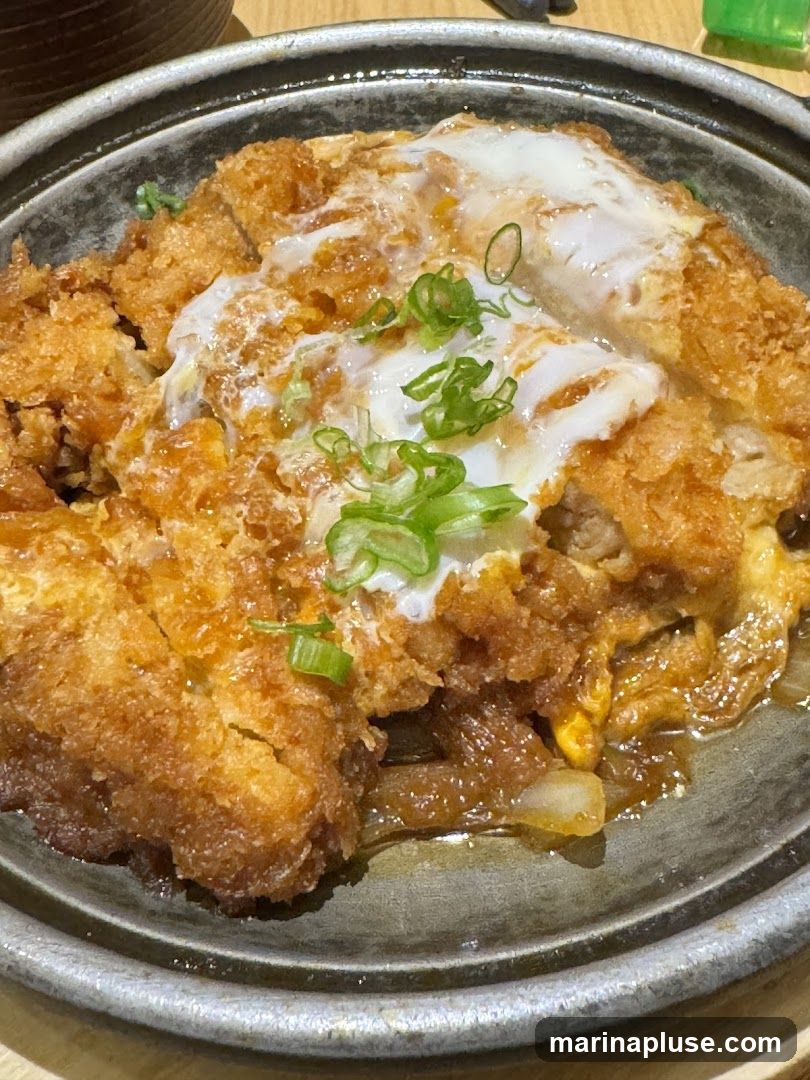 A close-up shot of a katsudon dish, featuring crispy fried pork cutlet simmered with onions and egg, served in a bowl. The dish is garnished with chopped green onions.