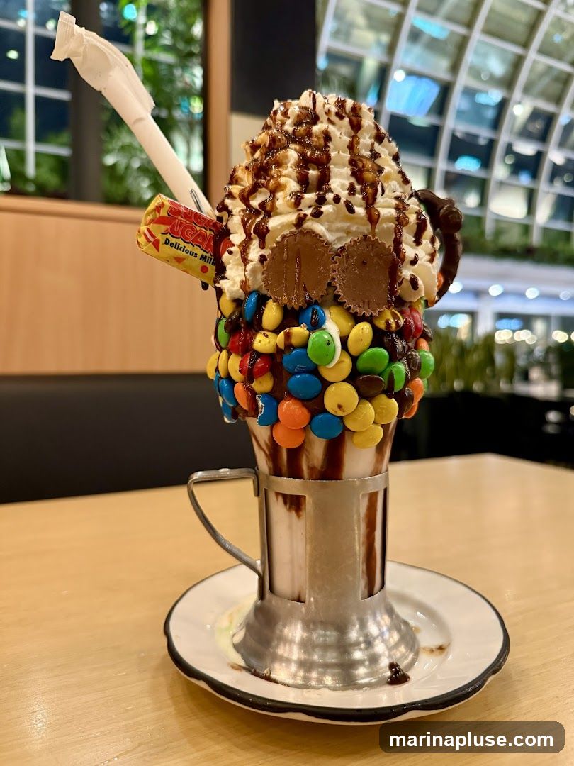A tall, elaborate milkshake topped with whipped cream, chocolate drizzle, M&Ms, peanut butter cups, and a candy bar. The milkshake is served in a metal holder on a saucer. The background shows a blurred interior with a modern, curved ceiling structure.
