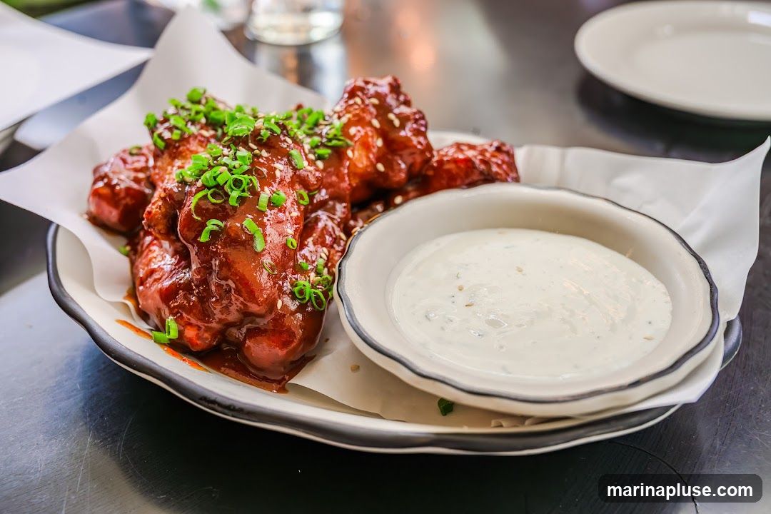 A plate of glossy, dark red chicken wings, sprinkled with sesame seeds and chopped green onions, is served with a side of creamy white dipping sauce. The wings are arranged on white parchment paper on a dark-rimmed plate. A white plate is visible in the blurred background.