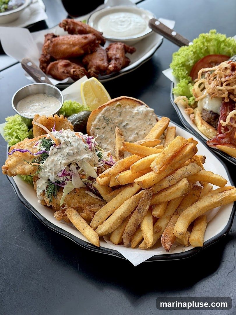 A close-up shot of a meal featuring fish and chips, a generous portion of french fries, and a side of chicken wings. The fish is battered and served with tartar sauce and coleslaw. The fries are golden brown and seasoned. The chicken wings are also golden brown and served with a dipping sauce. In the background, a burger with lettuce, tomato, and crispy onions is visible.