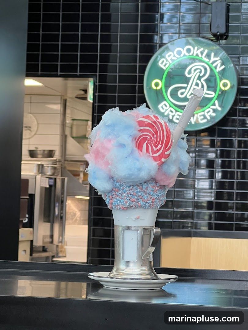 A tall glass filled with a white beverage is topped with a generous amount of pink and blue cotton candy. The rim of the glass is decorated with colorful sprinkles. A red and white swirled lollipop is stuck into the cotton candy, and a white straw is also visible. In the background, a neon sign reads "BROOKLYN BREWERY" in green lettering. The background also shows a tiled wall and a glimpse of a kitchen area.