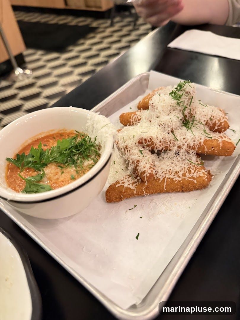 A metal tray holds several fried mozzarella sticks generously topped with shredded parmesan cheese and fresh herbs. To the left, a white bowl contains marinara sauce, also garnished with herbs and a sprinkle of cheese. The food is presented on a white paper liner within the tray. The background shows a patterned floor and a glimpse of a table setting.