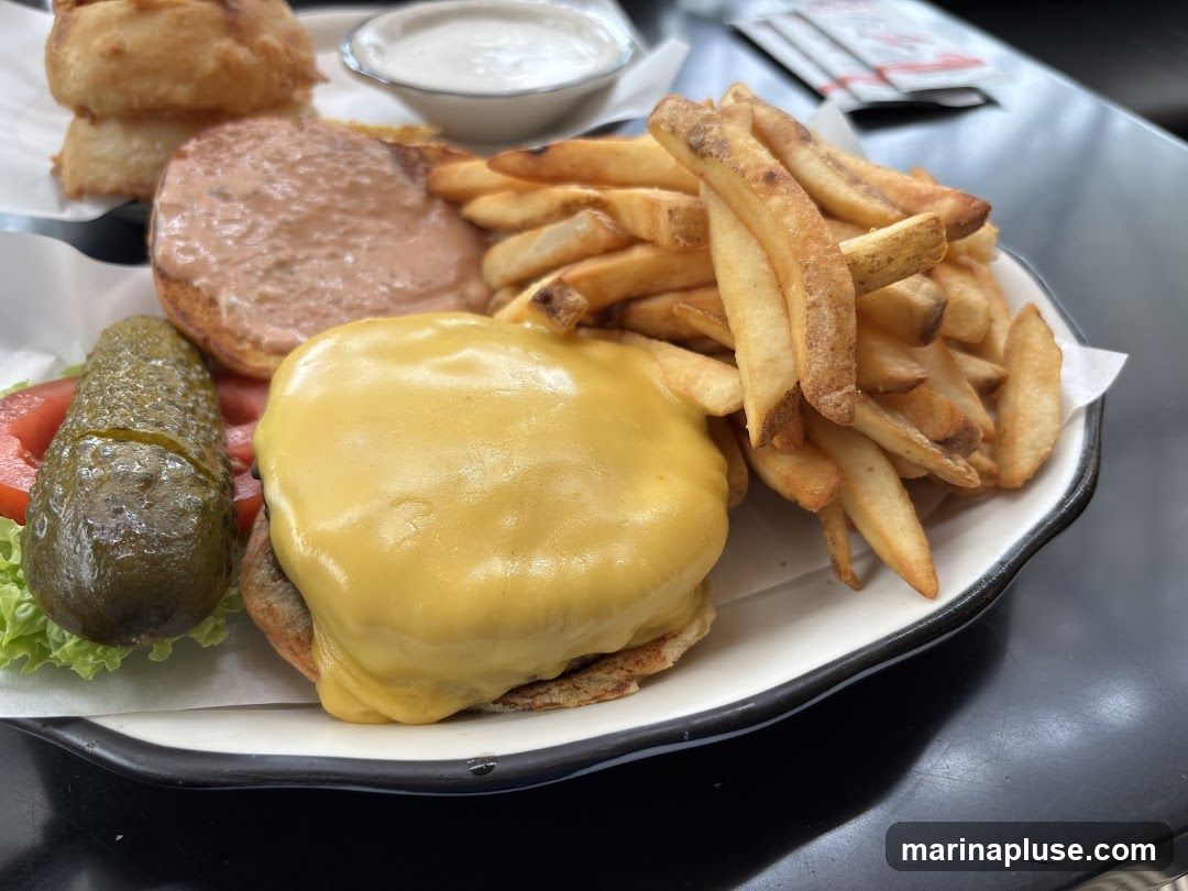 A close-up shot of a cheeseburger with melted yellow cheese, served with a side of french fries, a pickle spear, lettuce, and tomato. A small dish of white sauce and onion rings are visible in the background.
