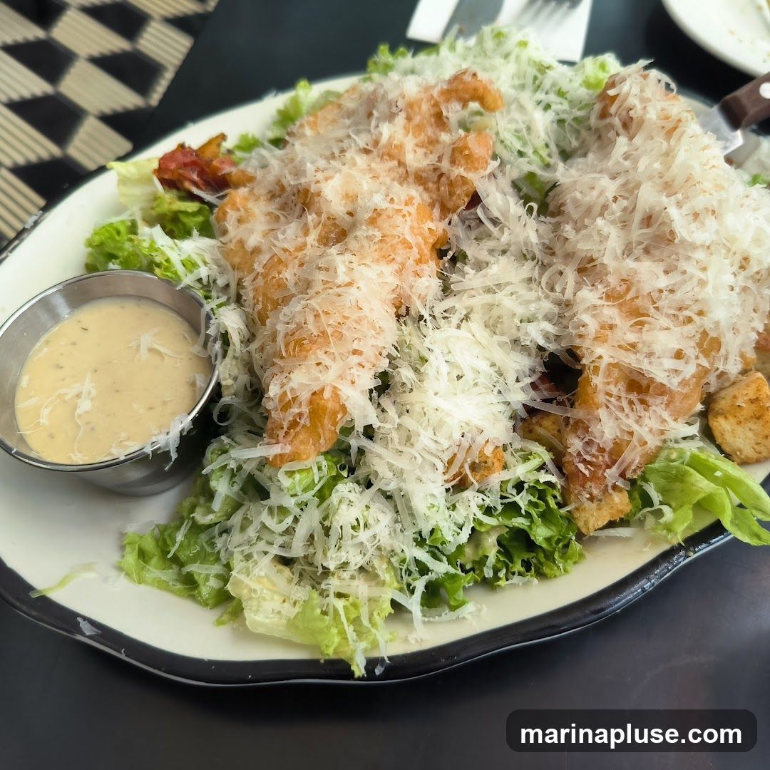 A close-up shot of a large chicken Caesar salad. The salad is served on a white plate with a dark rim. It features a bed of crisp romaine lettuce topped with generous portions of fried chicken tenders, shredded Parmesan cheese, croutons, and bacon bits. A small metal cup of creamy dressing is on the side. The background shows a black and white checkered pattern on a table.