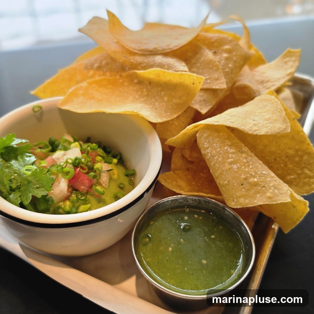 A close-up shot of a bowl of guacamole topped with chopped tomatoes, onions, and green onions, served with a side of green salsa and a large pile of tortilla chips. The food is presented on a metal tray.