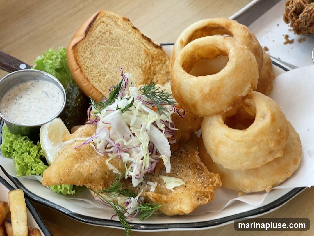 A plate of battered fish, coleslaw, a pickle, a lemon wedge, a small cup of sauce, a toasted bun, and several onion rings.