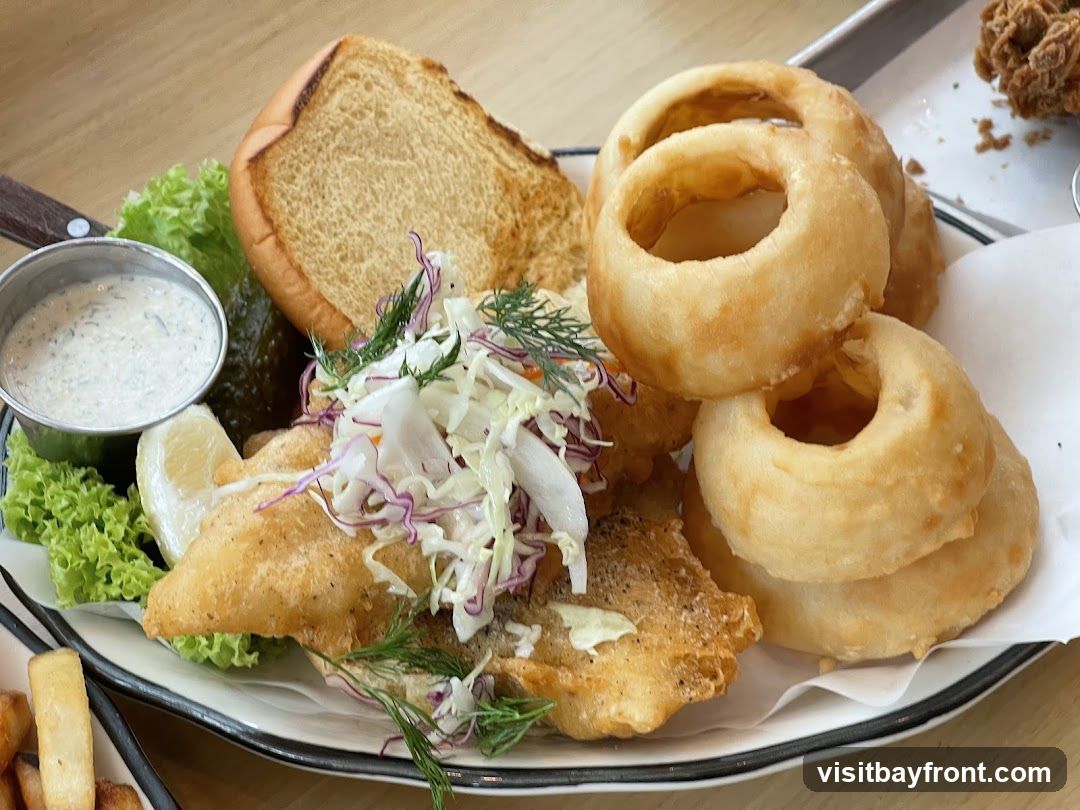 A plate of battered fish, coleslaw, a pickle, a lemon wedge, a small cup of sauce, a toasted bun, and several onion rings.