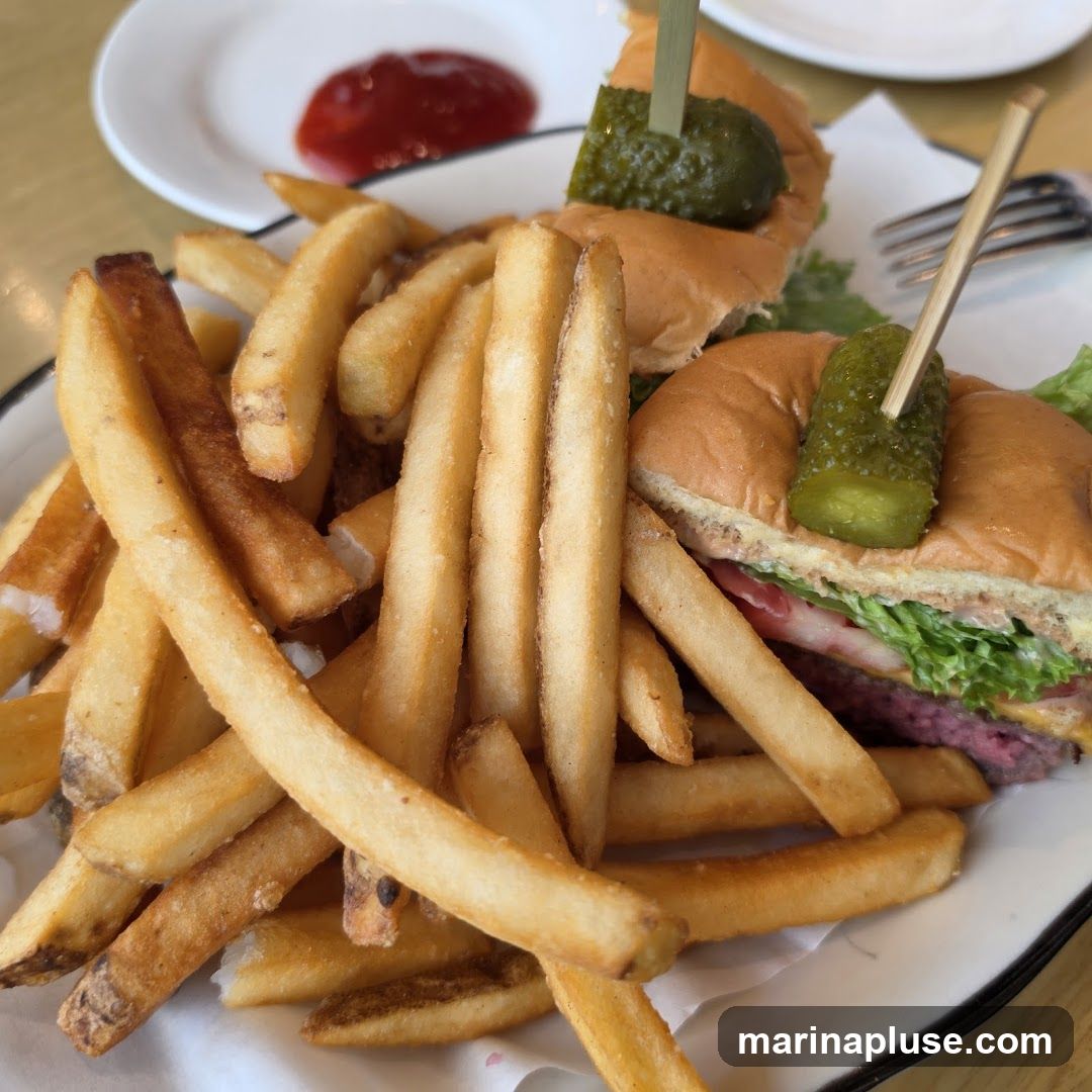 A close-up shot of two mini burgers served with a large portion of french fries. Each burger is topped with a pickle spear. A small dish of ketchup is visible in the background.