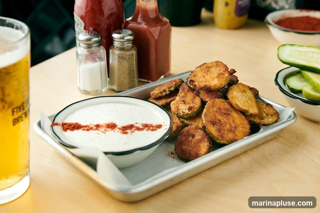 A metal tray holds a serving of fried pickles and a bowl of creamy dip, garnished with paprika. A glass of beer, salt and pepper shakers, and bottles of hot sauce are visible on a wooden table. Sliced cucumbers are in a small bowl in the background.
