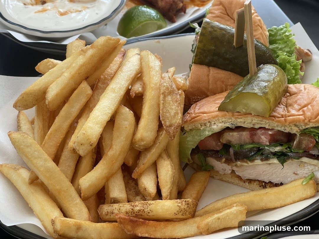 A close-up shot of a chicken sandwich served with a large portion of french fries. The sandwich features a toasted bun, lettuce, tomato, cheese, and a thick chicken breast. Two large pickle spears are skewered on top of the sandwich. In the background, a small bowl of dip and a lime wedge are visible.