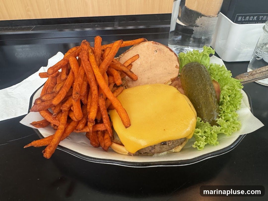 A close-up shot of a cheeseburger served with a side of sweet potato fries. The burger has a thick patty, melted yellow cheese, lettuce, and a dill pickle spear. The sweet potato fries are piled high next to the burger. A menu holder with the text "BLACK TAVERN CRAFT BURGERS & BEER" is visible in the background.