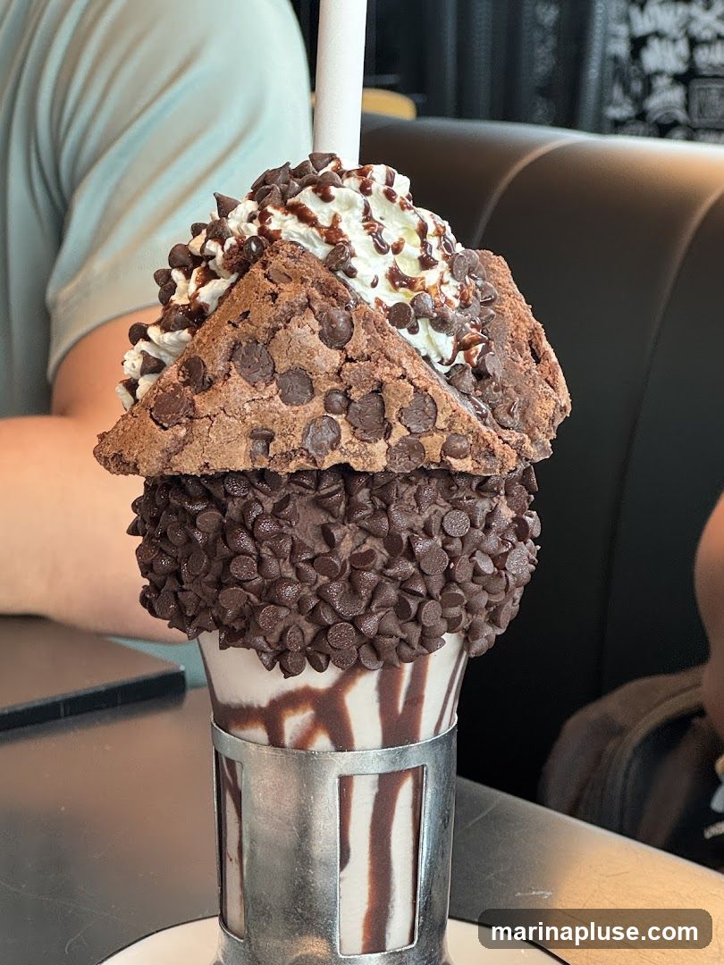A decadent milkshake topped with whipped cream, chocolate drizzle, a brownie, and a generous coating of mini chocolate chips. The glass is rimmed with chocolate chips and has chocolate syrup drizzled down the sides. It is served in a metal holder.