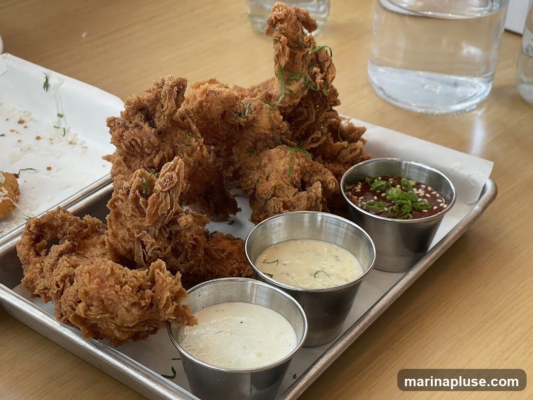 A close-up shot of a platter of crispy fried chicken pieces served with three dipping sauces in small metal cups. The chicken is golden brown and appears to be seasoned with herbs. One sauce is dark brown with sesame seeds and chopped green onions, another is a creamy white sauce, and the third is a lighter colored creamy sauce. The platter is on a light-colored wooden table, and glasses of water are visible in the background.