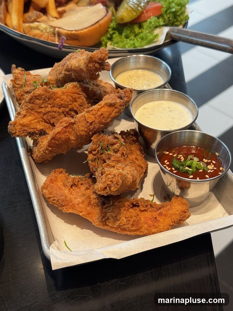 A close-up shot of a serving of crispy fried chicken strips arranged on a metal tray lined with white parchment paper. Three small metal cups hold dipping sauces: one appears to be a creamy white sauce, another a lighter colored sauce, and the third a reddish-brown sauce garnished with sesame seeds and chopped green onions. In the background, out of focus, is a burger with lettuce, tomato, and a pickle, along with a side of french fries. The table surface has a black and white checkered pattern.