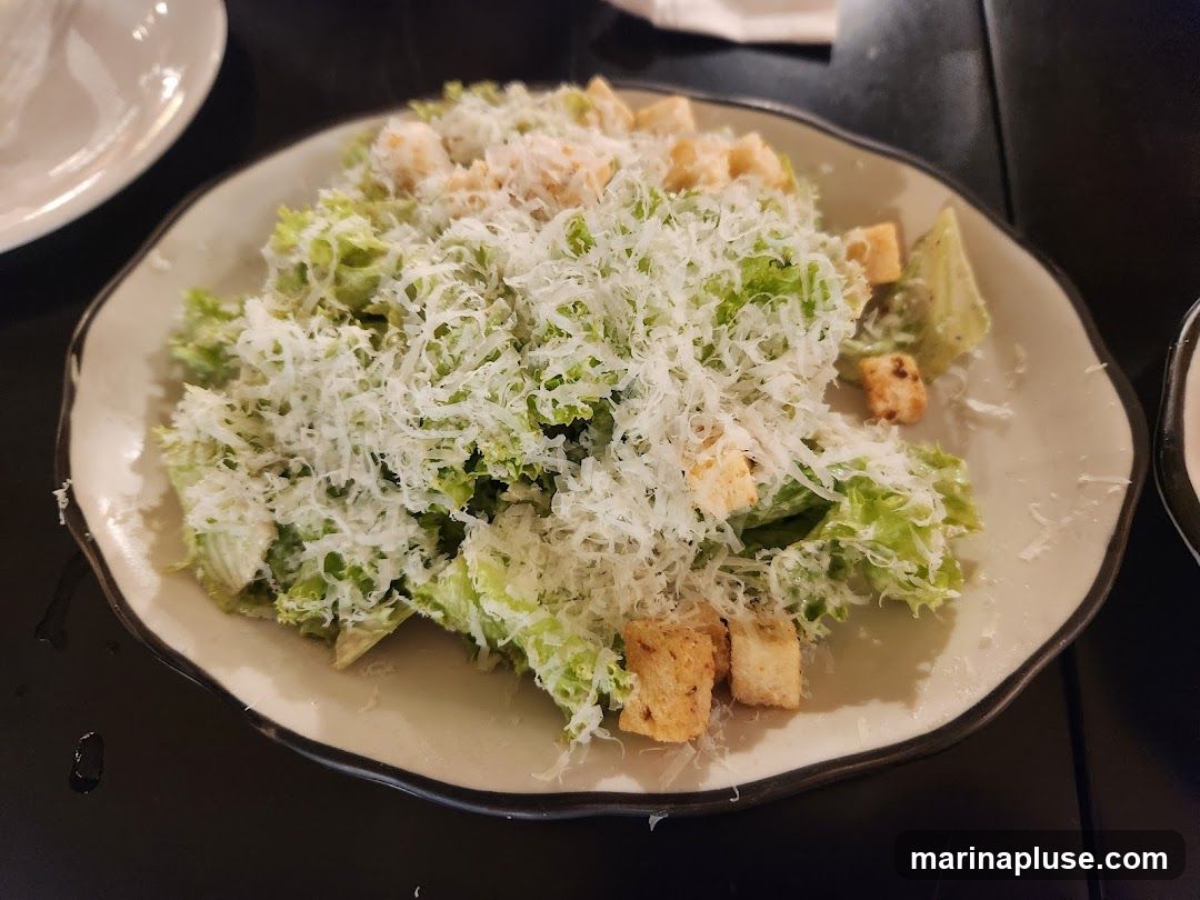 A close-up shot of a Caesar salad served on a light-colored, wavy-edged plate. The salad is made with crisp lettuce, croutons, and is generously topped with shredded Parmesan cheese and dressing. The background is dark, suggesting a restaurant setting.