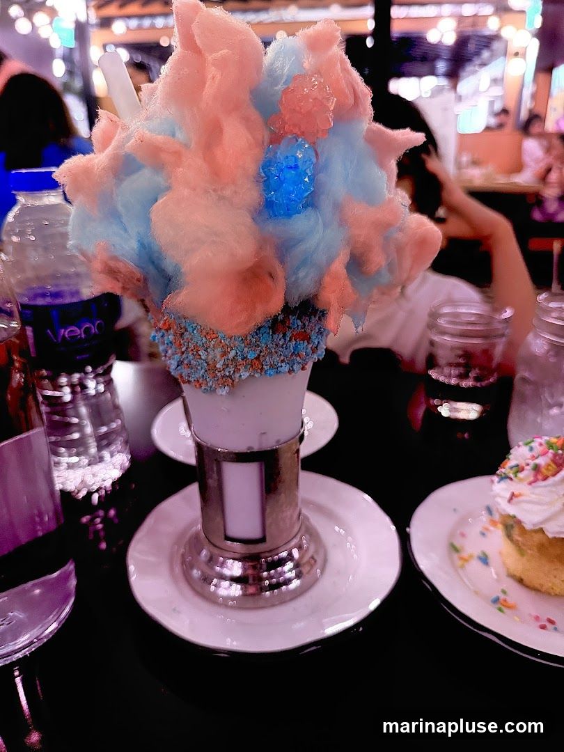 A tall glass filled with a milkshake or dessert, topped with blue and pink cotton candy and rock candy. The rim of the glass is coated with blue and pink sprinkles. A small cupcake with white frosting and sprinkles is on a plate to the right. A bottle of water and other glasses are on the table. The background is blurred, showing lights and people.