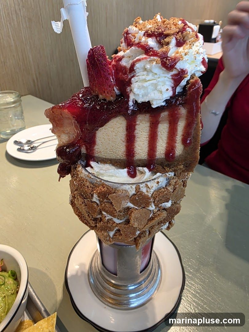 A decadent milkshake topped with a slice of cheesecake, whipped cream, strawberry sauce, and cookie crumbles. A strawberry slice garnishes the cheesecake. The milkshake glass is rimmed with cookie pieces. In the background, a person in a red shirt is visible, along with a glass of water and silverware on a plate. A small bowl of guacamole and chips is in the lower left corner.