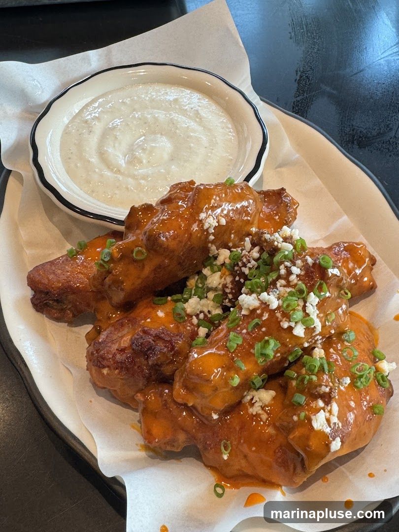 A plate of buffalo chicken wings coated in a glossy orange sauce, sprinkled with crumbled blue cheese and chopped green onions. A small bowl of creamy white dipping sauce is placed behind the wings. The wings are served on a white paper liner on a dark plate.