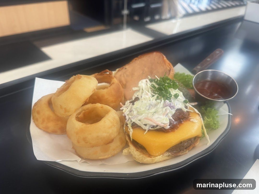 A close-up shot of a burger served with a side of onion rings and a small cup of barbecue sauce. The burger is topped with melted cheese, coleslaw, and chives. A slice of meat and lettuce are also visible on the plate.