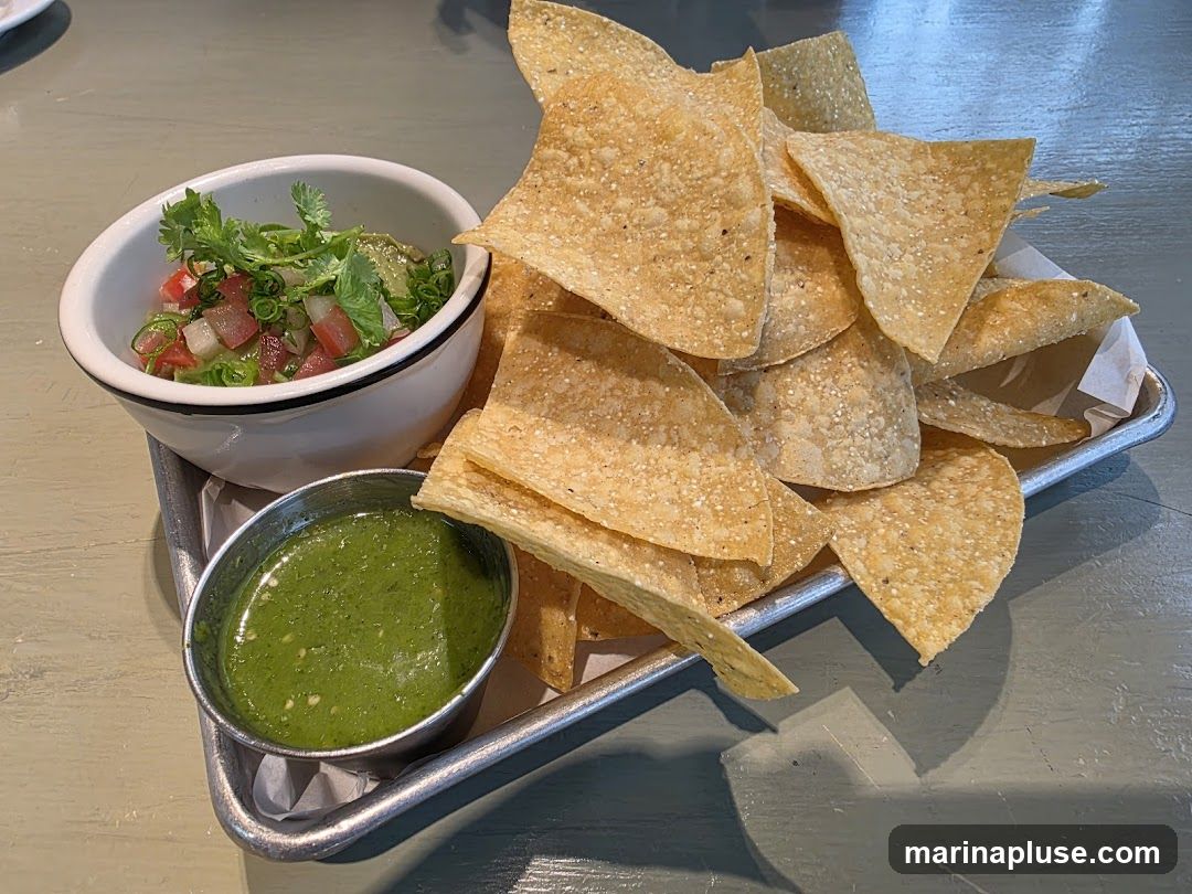 A metal tray holds a white bowl of pico de gallo and a small metal cup of green salsa, accompanied by a pile of tortilla chips.