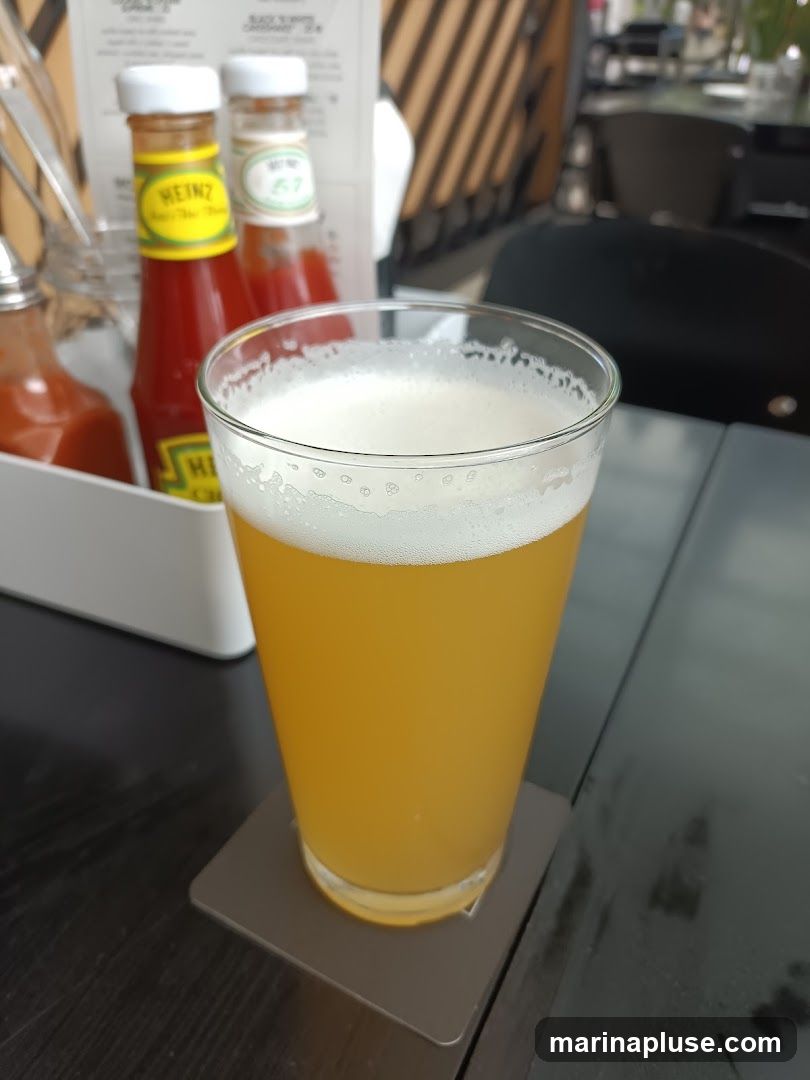 A glass of beer with a foamy head sits on a coaster on a dark table. In the background, out of focus, are bottles of Heinz ketchup and another condiment, along with cutlery in a holder. A menu is also visible behind the condiments.