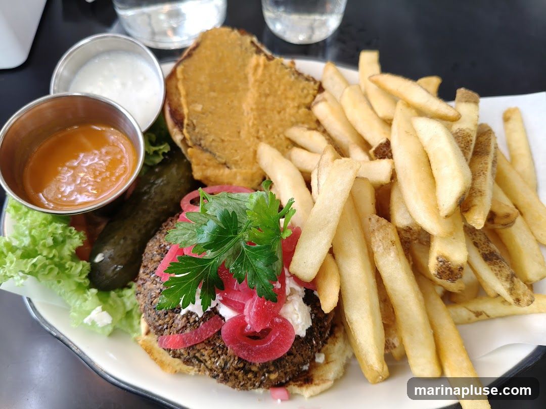 A close-up shot of a veggie burger served with a side of french fries, lettuce, a pickle, and two dipping sauces. The burger patty appears to be made of grains and vegetables, topped with white cheese, pickled red onions, and fresh parsley. One sauce is a creamy white, and the other is a reddish-orange sauce. The fries are golden brown and crispy.