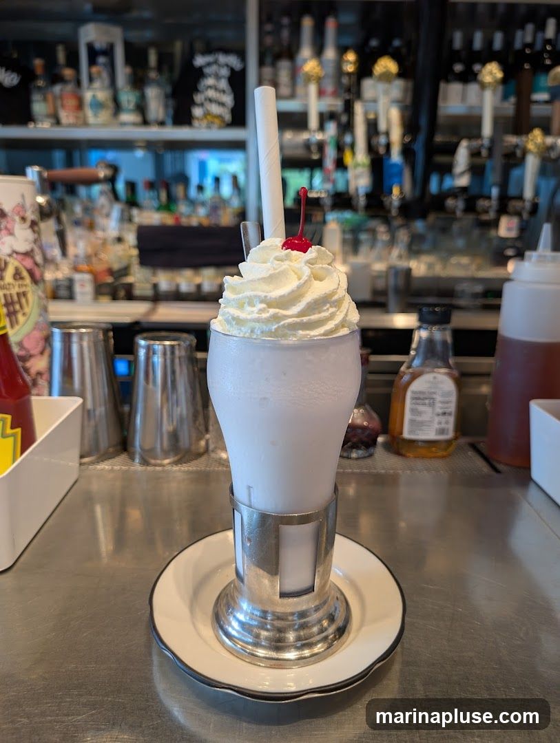 A close-up shot of a vanilla milkshake topped with whipped cream and a maraschino cherry, served in a glass with a metal holder and saucer. The background shows a bar with bottles of alcohol and taps.