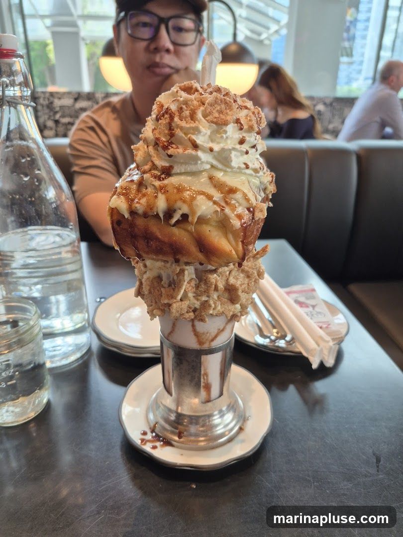 A close-up shot of a decadent dessert served in a metallic holder. The dessert is topped with a generous swirl of whipped cream, drizzled with caramel sauce, and sprinkled with a crumble topping. A person wearing glasses and a cap is visible in the background, out of focus. Other patrons and booth seating are also visible in the blurred background, suggesting a restaurant or cafe setting.