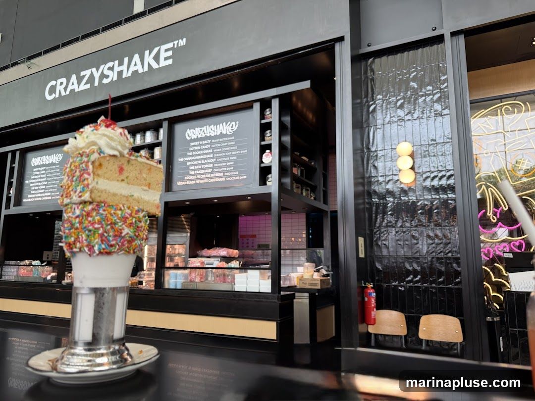 A close-up of a "Crazy Shake" milkshake topped with a slice of cake and sprinkles, displayed in front of the Crazy Shake shop counter. The shop's signage and menu boards are visible in the background.