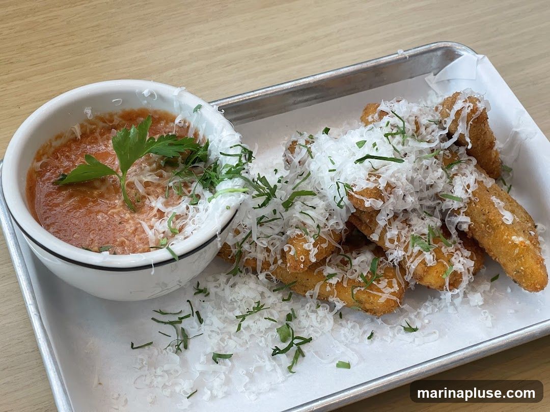A serving of fried mozzarella sticks, generously topped with shredded Parmesan cheese and fresh herbs, is presented on a metal tray lined with white parchment paper. A small white bowl filled with marinara sauce, garnished with a sprig of parsley and more Parmesan cheese, sits to the left of the mozzarella sticks. The food is arranged on a light-colored wooden surface.