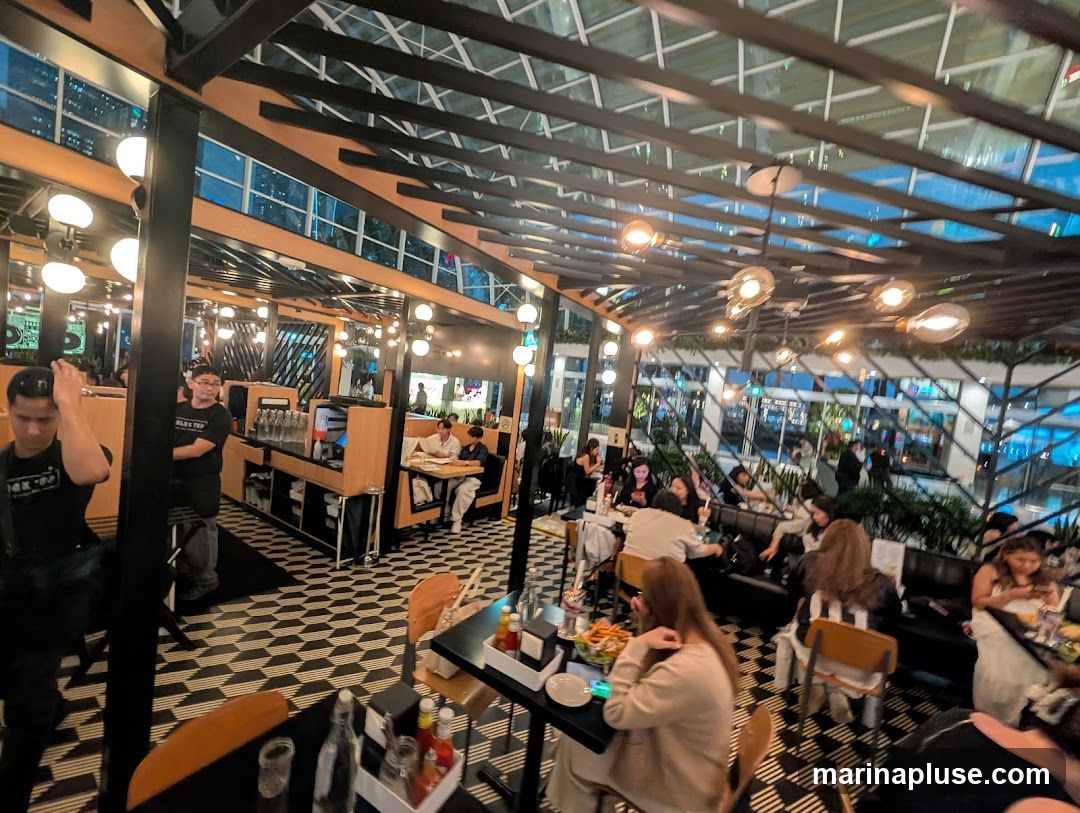 A busy restaurant interior with patrons seated at tables and booths. The space features a modern design with a black and white geometric tiled floor, wooden accents, and a glass ceiling with exposed beams. Numerous Edison-style light bulbs hang from the ceiling, providing warm illumination. Staff members are visible behind a counter area.