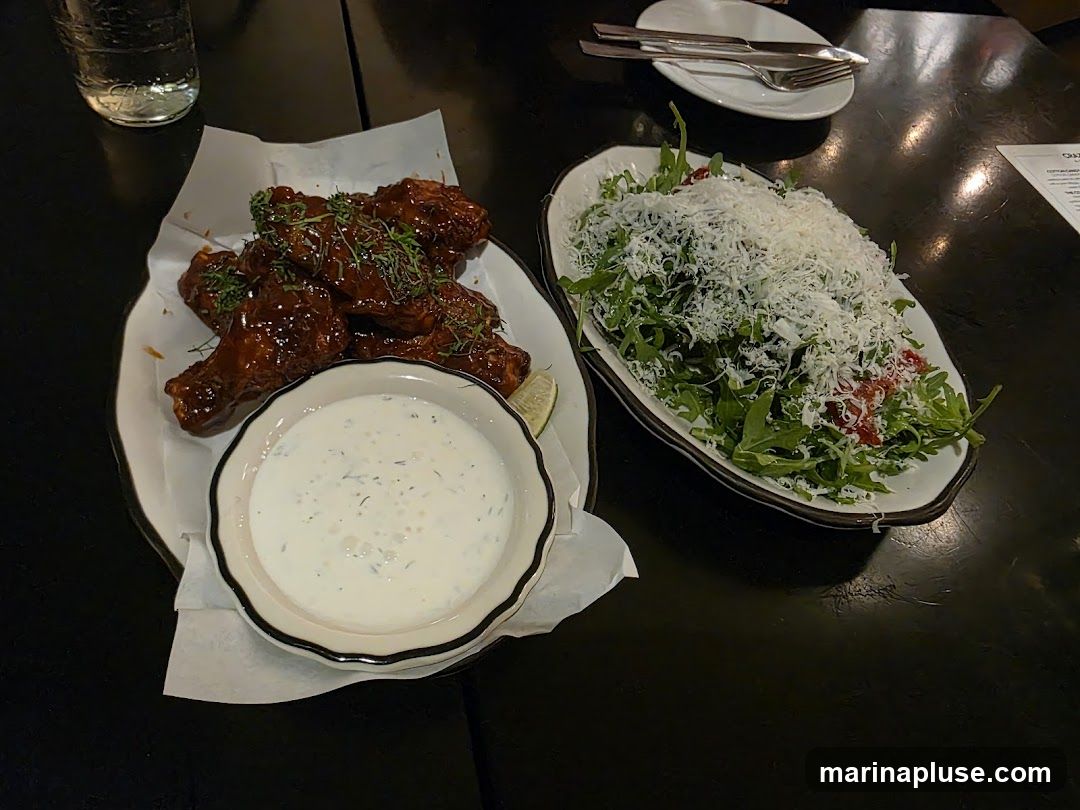 A plate of glossy, dark red chicken wings with green herbs sprinkled on top sits next to a bowl of creamy dipping sauce. To the right, a white oval dish holds a large salad composed of arugula, cherry tomatoes, and a generous topping of shredded white cheese. A glass of water and cutlery on a small plate are also visible on the dark table. A corner of a menu is visible on the right side of the image.