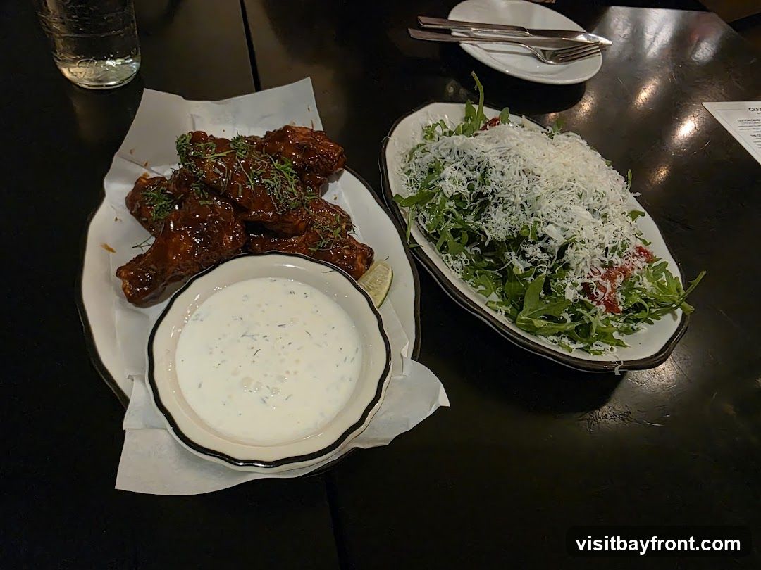 A plate of glossy, dark red chicken wings with green herbs sprinkled on top sits next to a bowl of creamy dipping sauce. To the right, a white oval dish holds a large salad composed of arugula, cherry tomatoes, and a generous topping of shredded white cheese. A glass of water and cutlery on a small plate are also visible on the dark table. A corner of a menu is visible on the right side of the image.