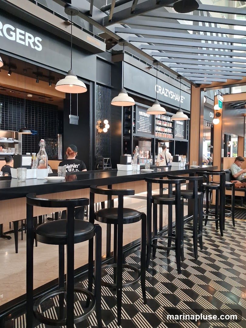 The image shows the interior of a modern burger restaurant. A long, black counter with bar stools is in the foreground, with a black and white geometric tiled floor. Behind the counter, a black wall features signage that reads "BURGERS" and "CRAZYSHAKE". Several pendant lights hang from the ceiling. Shelves stocked with jars and bottles are visible behind the counter, along with a menu board. Staff members are seen working behind the counter, and customers are seated at tables in the background.