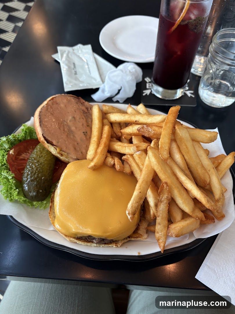 A close-up view of a cheeseburger with a melted cheese slice, served with a side of french fries. The burger is open-faced, revealing the patty and bun. Lettuce, tomato, and a pickle spear are visible on the side. A glass of red sangria and a mason jar of water are also on the table, along with a wet wipe packet and a crumpled napkin.