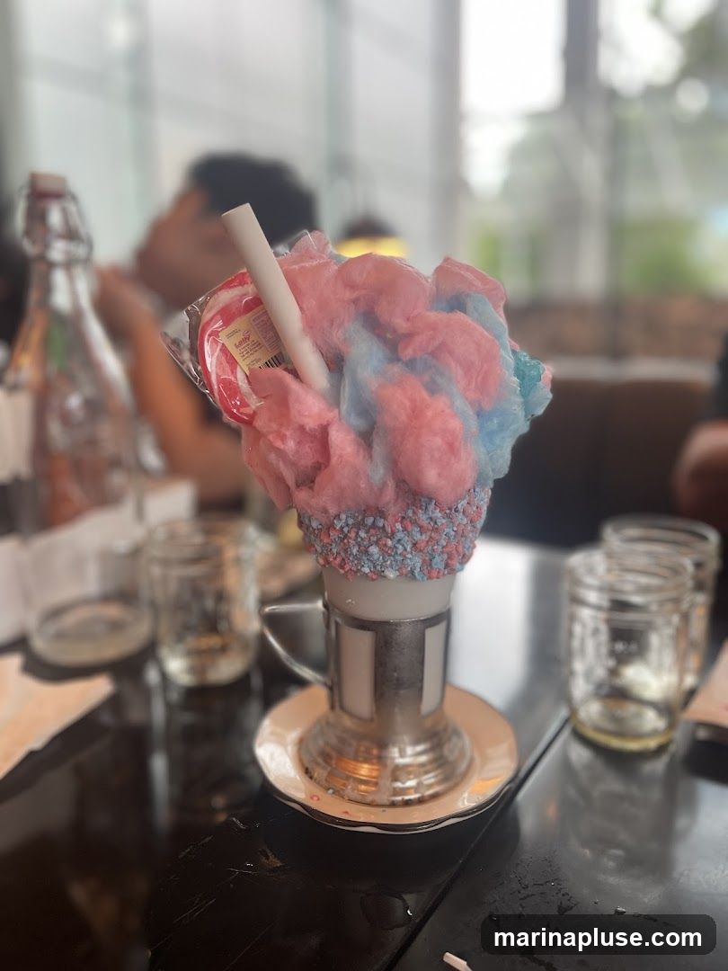 A close-up shot of a decadent dessert, possibly a milkshake or sundae, topped with fluffy pink and blue cotton candy. A red and white swirled lollipop is inserted into the dessert, along with a white straw. The rim of the dessert cup is coated in colorful sprinkles. The dessert sits on a saucer on a dark, reflective table. In the background, out of focus, are glass jars and a person.