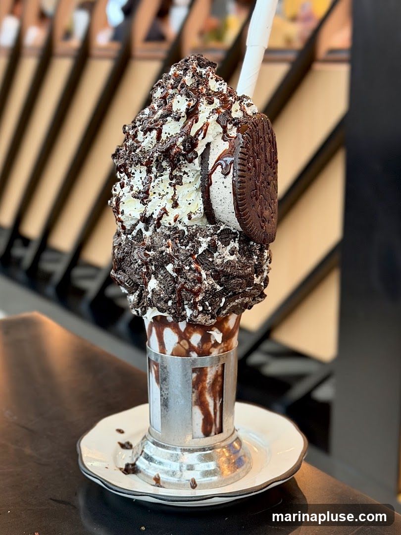 A tall glass filled with a thick milkshake, topped with whipped cream, chocolate syrup, crushed Oreos, and a whole Oreo cookie. The glass is served in a metallic holder on a white saucer. The background shows a blurred interior with wooden elements.