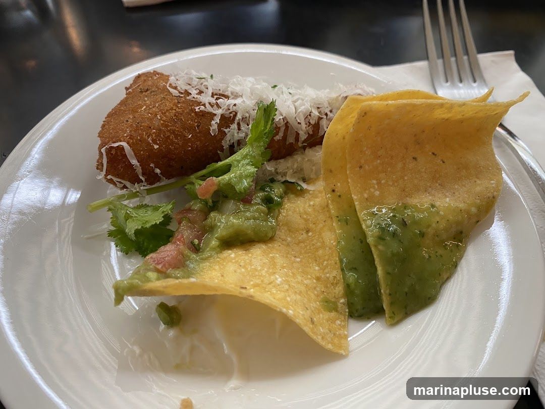 A white plate holds a fried, breaded item topped with shredded cheese and cilantro. Next to it are tortilla chips with guacamole and a green sauce. A fork and napkin are visible in the background.