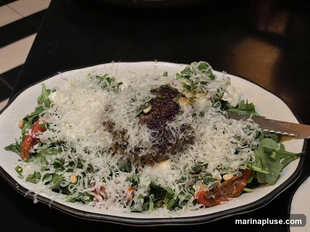 A close-up shot of a salad served in a white bowl. The salad contains arugula, cherry tomatoes, pine nuts, and is generously topped with shredded cheese. A dark, rich dressing or sauce is visible in the center, and a knife with the brand 'Royal Steel' is placed on the side of the bowl. The background is dark, with a black and white striped pattern visible on the left.
