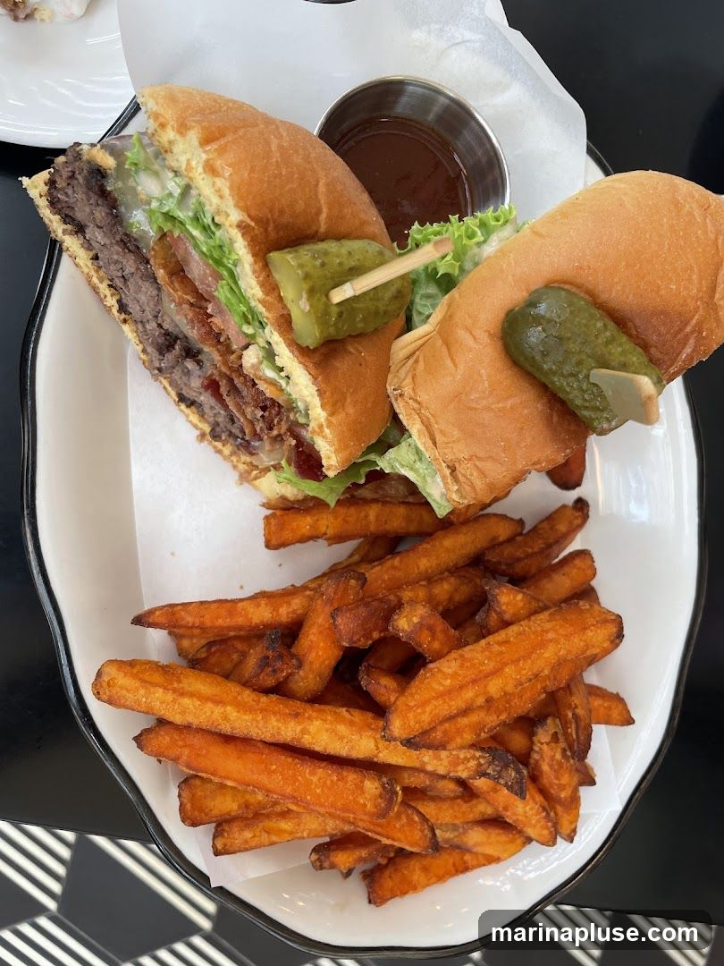 A close-up overhead shot of a gourmet burger cut in half, served with a side of sweet potato fries and a small metal cup of dipping sauce. The burger is on a bun with lettuce, cheese, and bacon. Two pickle spears are skewered on toothpicks, one in each half of the burger. The sweet potato fries are golden brown and piled high on the plate.