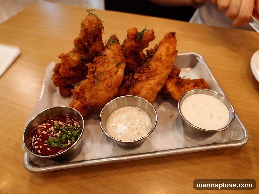 A metal tray holds several pieces of golden-brown fried chicken strips, garnished with fresh herbs. Three small metal cups contain dipping sauces: one with a dark red sauce topped with sesame seeds and chopped green onions, and two with creamy white sauces. The tray is placed on a wooden table.
