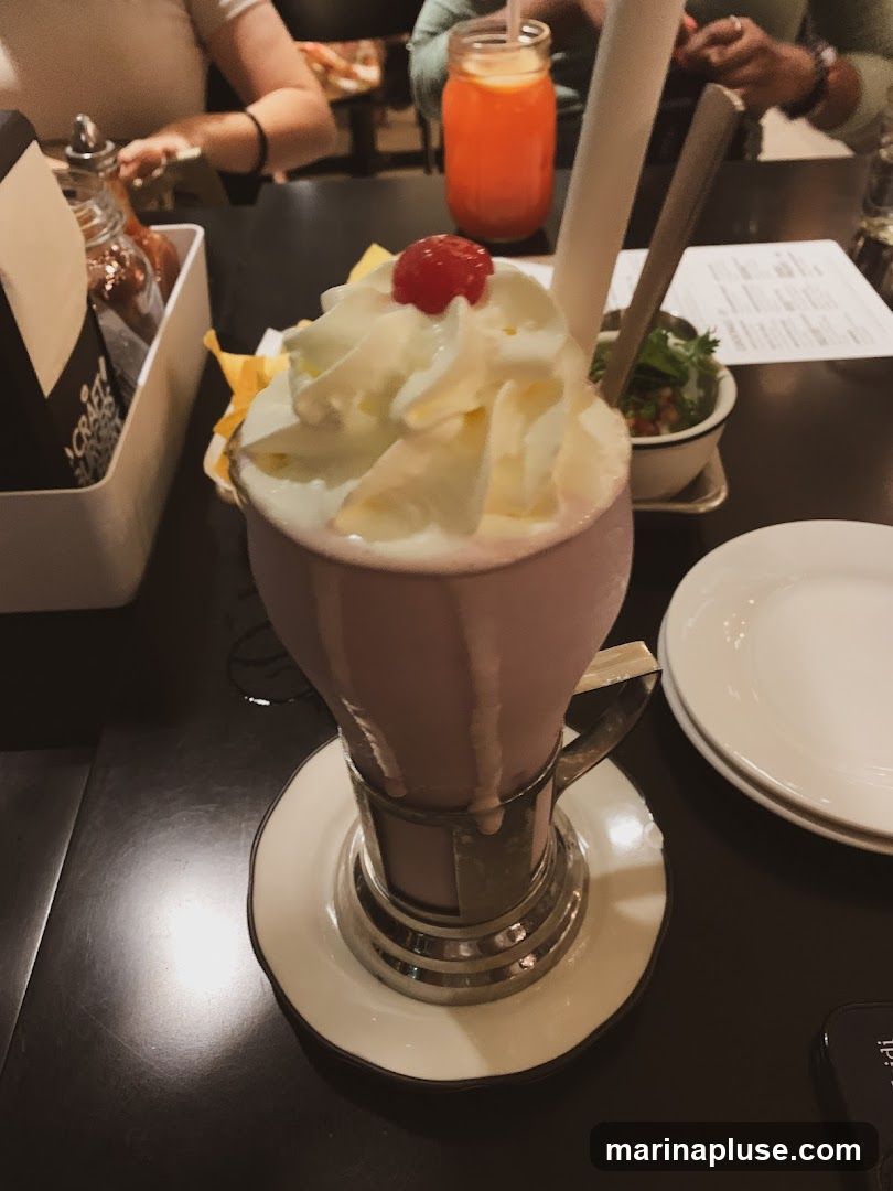A close-up of a pink milkshake topped with whipped cream and a cherry. The milkshake is served in a glass with a metal holder and a saucer. In the background, there are people, a drink in a mason jar, a small bowl of salsa or dip, and a menu on the table.