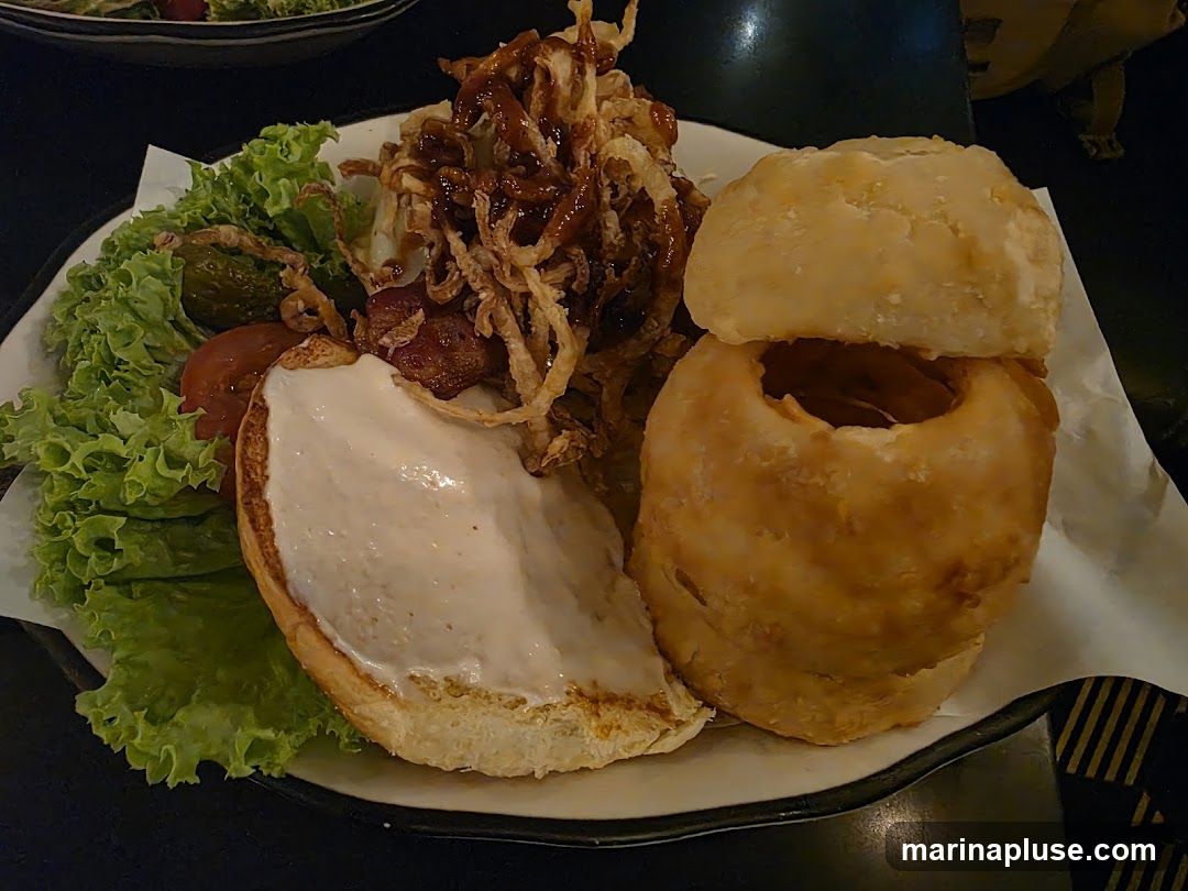 A deconstructed burger is served on a plate with a side of lettuce, tomato, pickles, and crispy onion rings. The burger bun is open, with one half spread with a creamy sauce. The other half of the bun is placed on top of a large, hollowed-out onion ring, which is stacked on another onion ring. The burger patty appears to be topped with barbecue sauce and crispy fried onions.