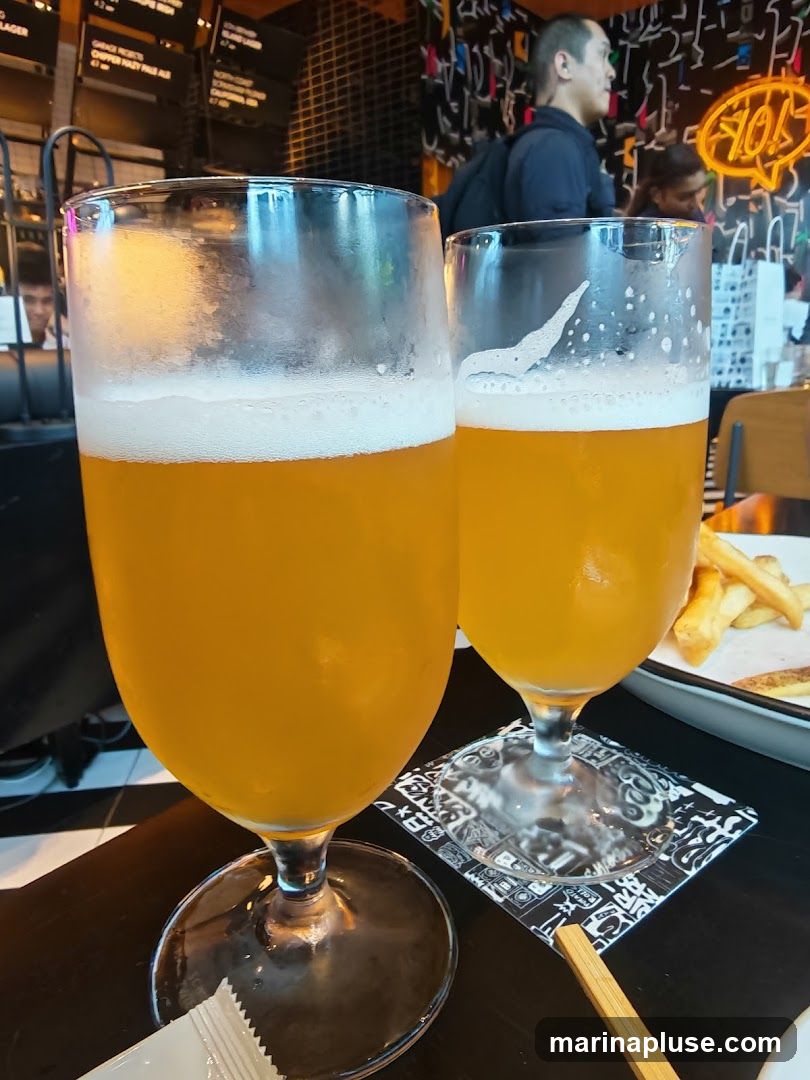 Two glasses of beer with frothy heads are on a table. In the background, a plate of french fries is visible, along with a neon sign that says "FIOH". People are also visible in the background.