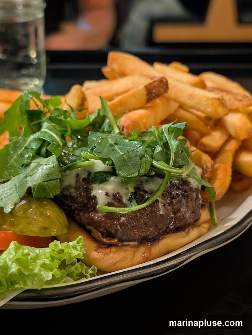 A close-up shot of a gourmet burger served with a side of golden french fries. The burger is topped with fresh arugula and a creamy sauce, with lettuce, tomato, and a pickle on the side. A glass of water is visible in the background.