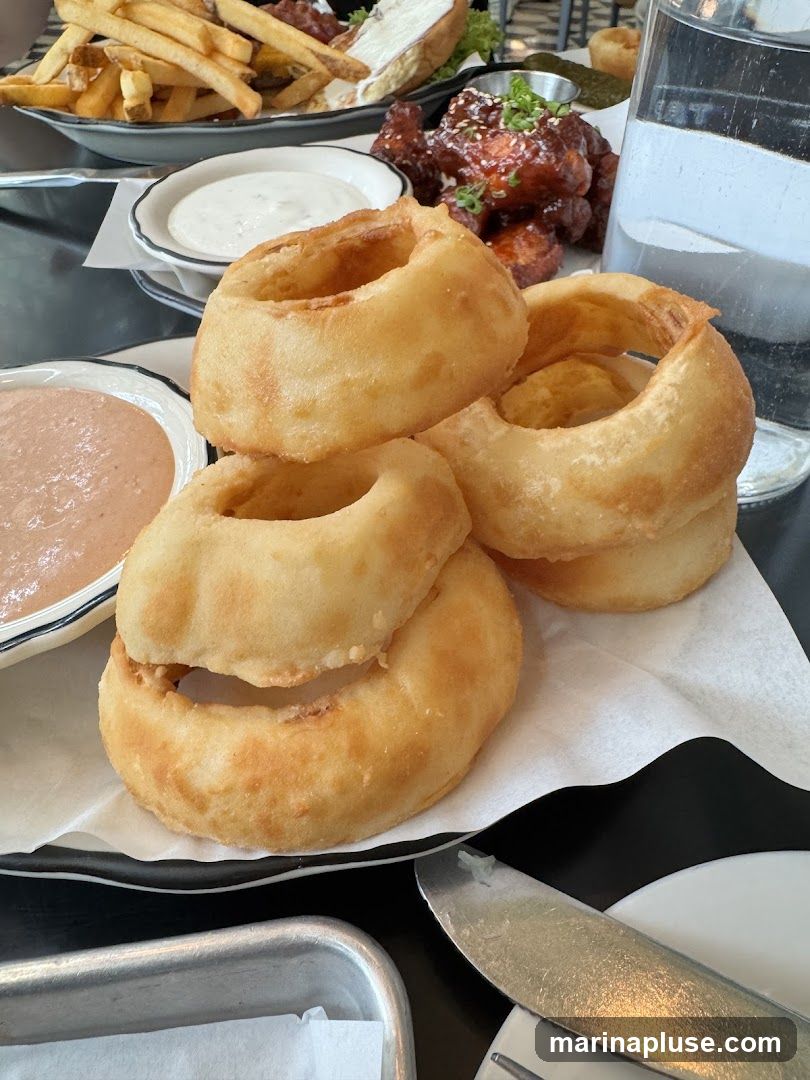 A close-up shot of a plate of golden-brown onion rings, stacked high. In the background, a platter of french fries, a dish of chicken wings with a glossy sauce, and a small bowl of dipping sauce are visible. A glass of water sits to the right.