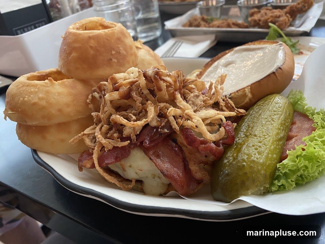 A close-up shot of a burger served with a side of onion rings. The burger is topped with crispy fried onions, bacon, and cheese, and is accompanied by a large pickle spear and lettuce. The bun is open, revealing a creamy sauce. In the background, other dishes and drinks are visible on tables.
