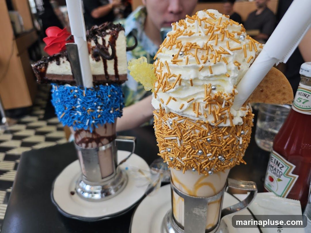Two elaborately decorated milkshakes are displayed on a table. The milkshake on the left is topped with blue sprinkles, chocolate cake slices, and a red rose. The milkshake on the right is covered in gold sprinkles and topped with whipped cream and a cookie. A bottle of Heinz ketchup is visible in the background.