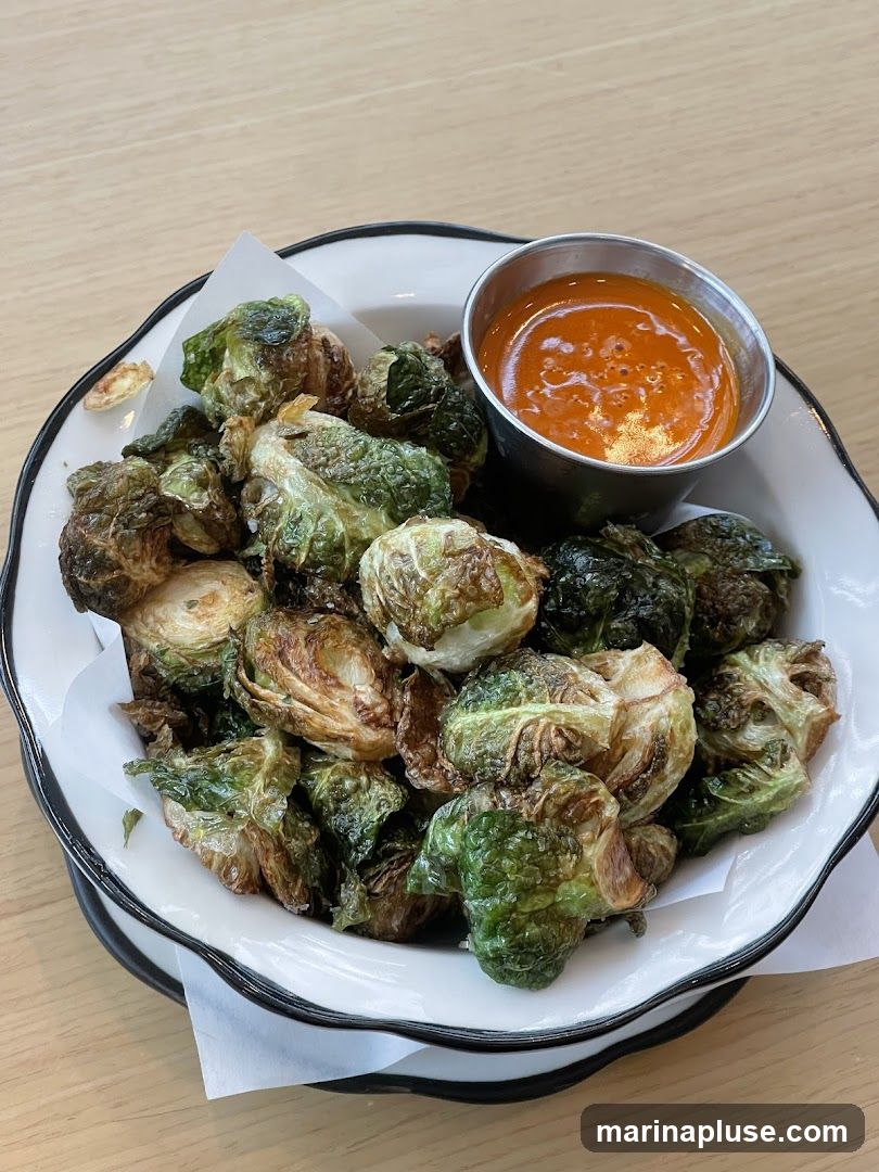 A bowl of crispy, roasted Brussels sprouts is served with a small metal cup of orange dipping sauce. The Brussels sprouts are a deep green and brown, indicating they are well-cooked and slightly charred. The dipping sauce appears to be a smooth, vibrant orange color. The dish is presented in a white bowl with a black rim, lined with white parchment paper. The setting appears to be a restaurant table with a light wood grain surface.