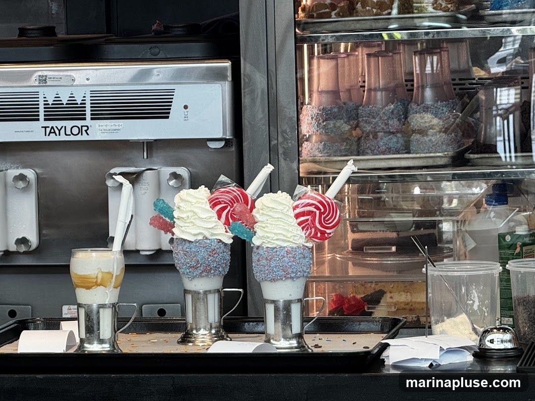 A display of elaborate desserts, including milkshakes topped with lollipops and rock candy, and a layered drink. In the background, a Taylor soft-serve machine is visible, along with a refrigerated display case filled with various cakes and pastries.